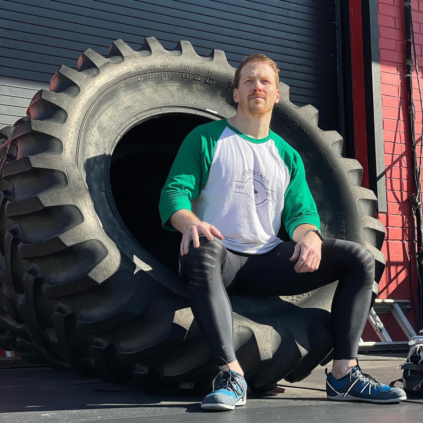 Man sitting on a large industrial tire wearing the Folding Chair Barbell Club raglan tee.
